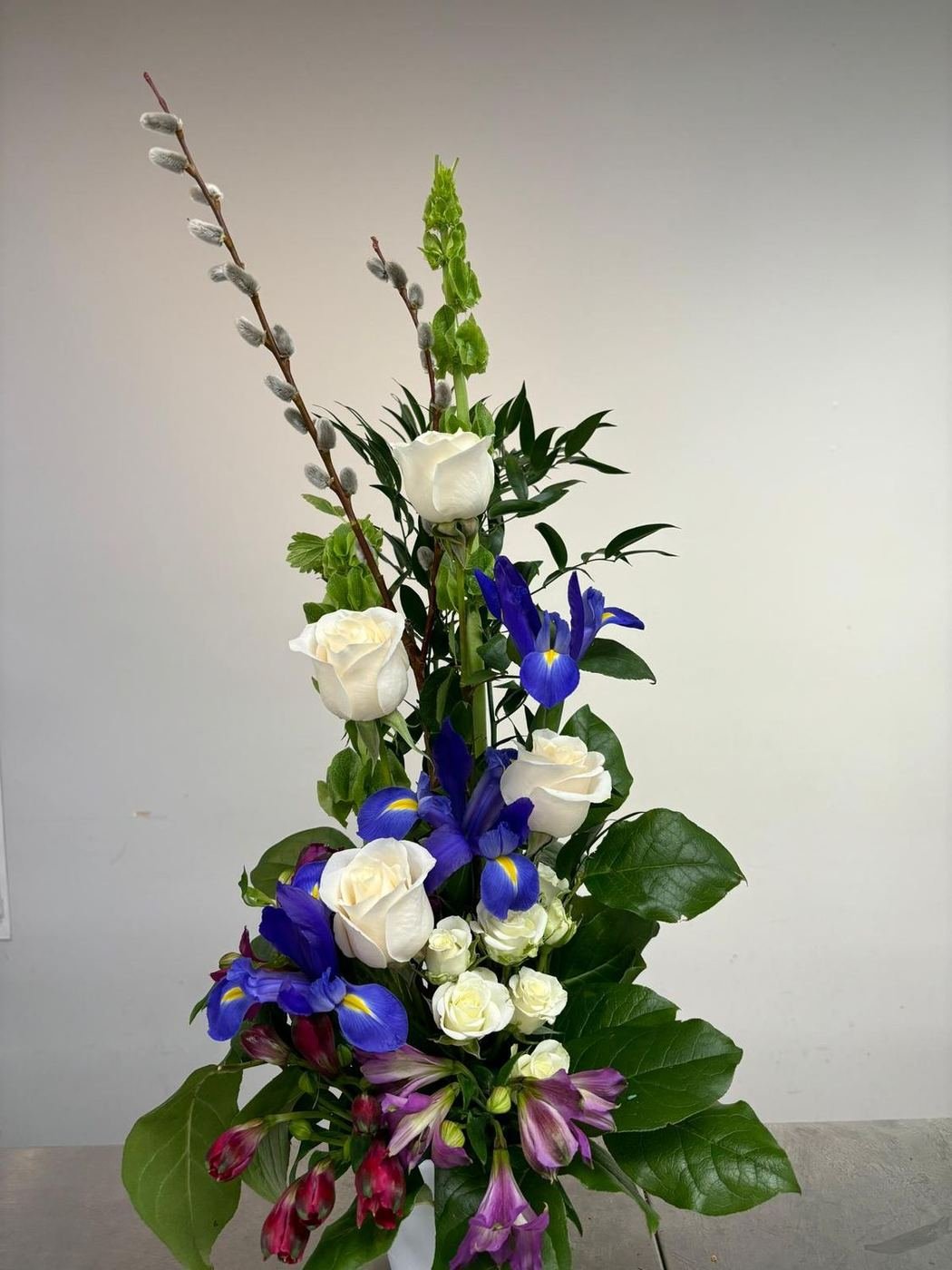 Tall arrangement of ivory roses, blue iris, bells of Ireland and pussy willow